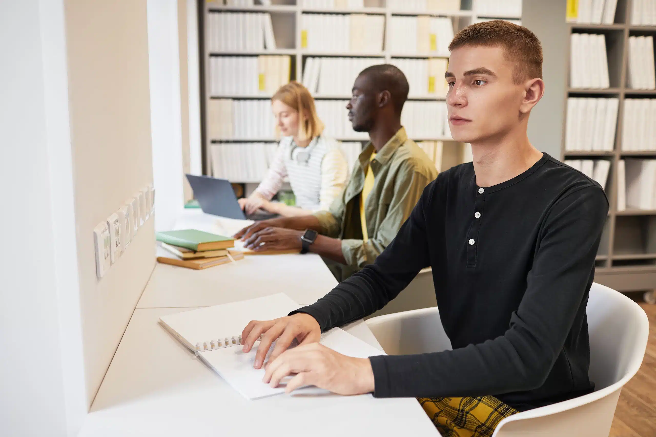 Synskadad person läser punktskrift intill två andra personer på bibliotek.