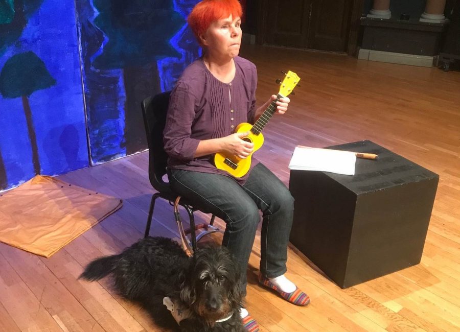 Sofia sitter på en stol med en gul ukulele i handen och en svart ledarhund ligger intill på golvet.
