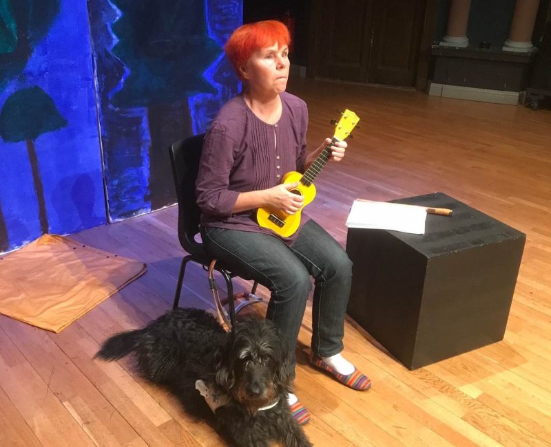 Sofia sitter på en stol med en gul ukulele i handen och en svart ledarhund ligger intill på golvet.